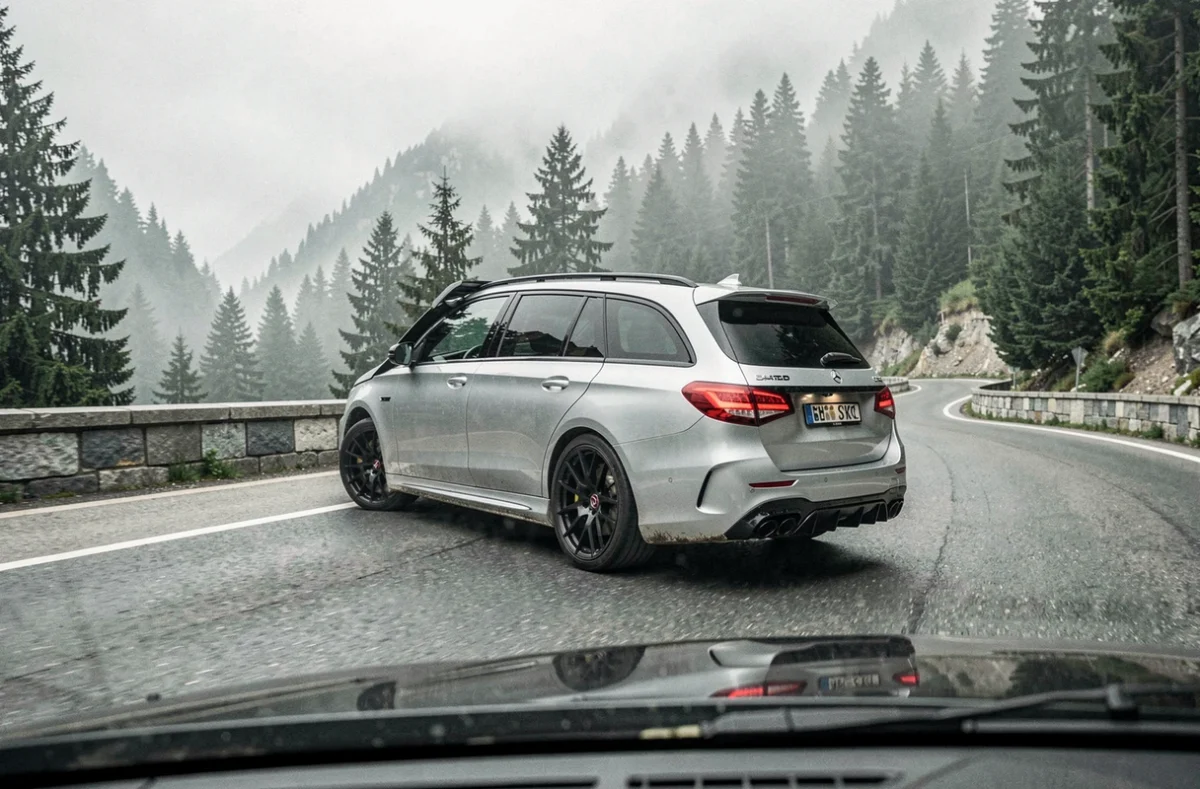 Brabus Mercedes E63 S Estate argentée en virage sur une route alpine brumeuse avec forêt de pins et barrières en pierre