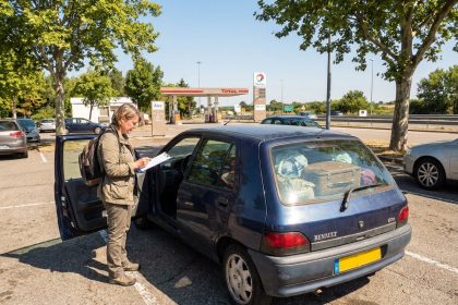 Une femme consulte une liste de vérification avant de partir en vacances avec une vieille citadine garée sur une aire d'autoroute